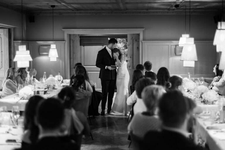 Braut und Bräutigam halten eine Rede vor dem Hochzeitsessen, umgeben von Gästen am Tisch