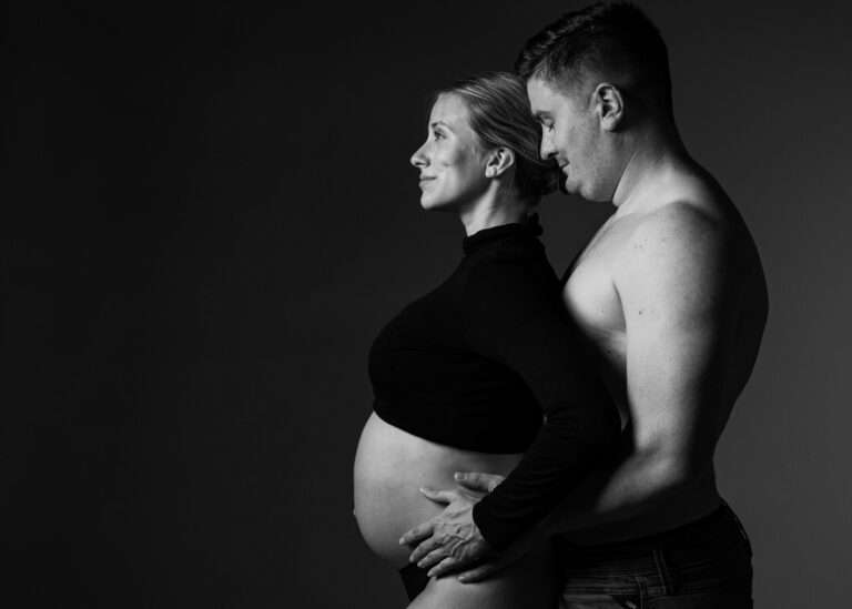 Fotografie Schwarzweiß-Babybauchfoto eines Paares im Profil. Der Partner hält den Babybauch der schwangeren Frau, emotionale Schwangerschaftsfotografie im Studio.